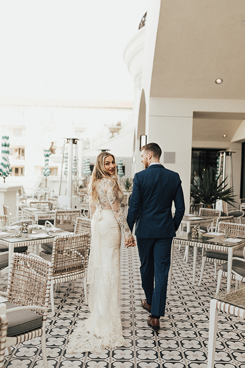 Dana point outdoor wedding shoot at monarch beach resort bride form fitting lace gown with long sleeves and high neckline with groom navy blue suit with matching vest and white dress shirt with long blush pink striped tie and matching pocket square walking and holding hands