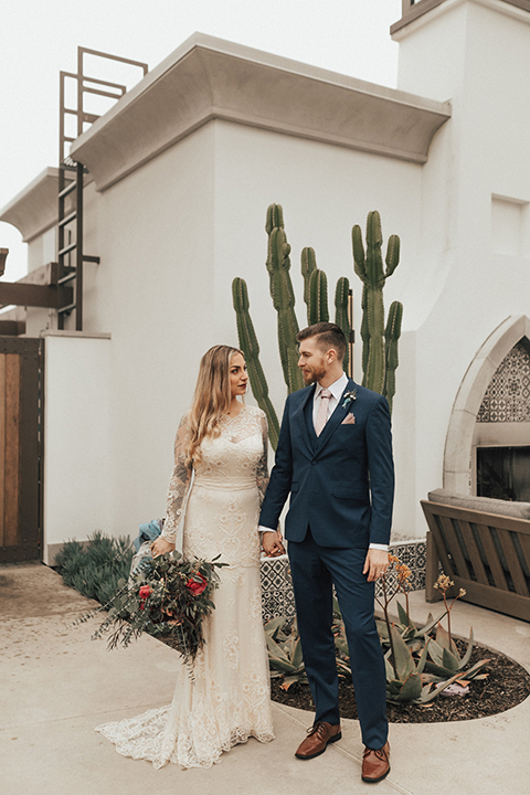 Dana point outdoor wedding shoot at monarch beach resort bride form fitting lace gown with long sleeves and high neckline with groom navy blue suit with matching vest and white dress shirt with long blush pink striped tie and matching pocket square holding hands