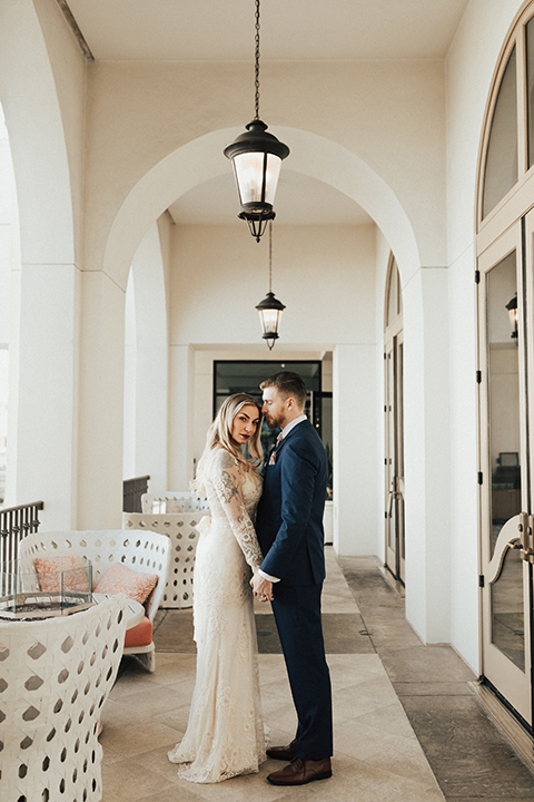 Dana point outdoor wedding shoot at monarch beach resort bride form fitting lace gown with long sleeves and high neckline with groom navy blue suit with matching vest and white dress shirt with long blush pink striped tie and matching pocket square holding hands and hugging