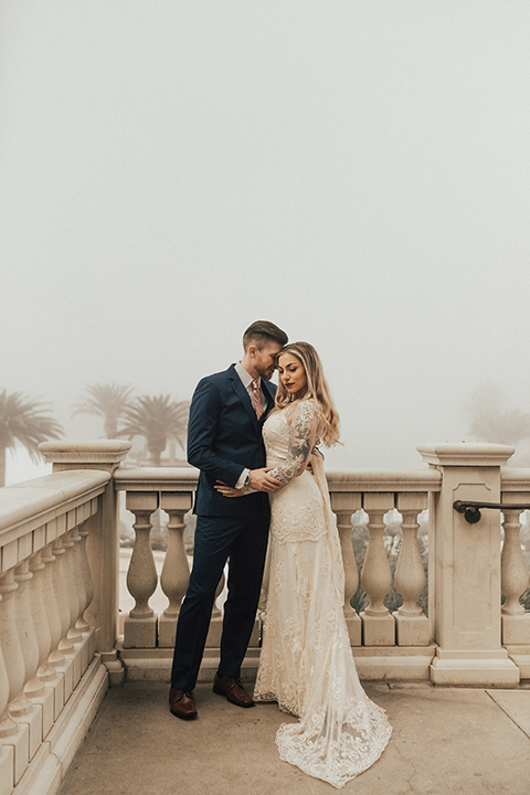 Dana point outdoor wedding shoot at monarch beach resort bride form fitting lace gown with long sleeves and high neckline with groom navy blue suit with matching vest and white dress shirt with long blush pink striped tie and matching pocket square hugging