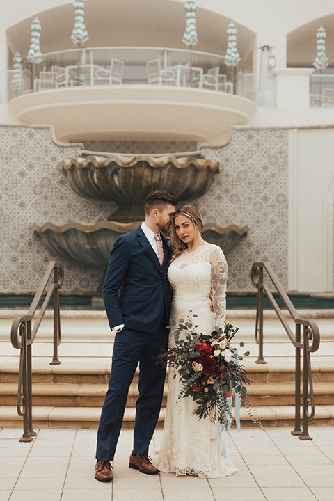 Dana point outdoor wedding shoot at monarch beach resort bride form fitting lace gown with long sleeves and high neckline with groom navy blue suit with matching vest and white dress shirt with long blush pink striped tie and matching pocket square bride holding red and green floral bridal bouquet
