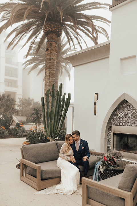 Dana point outdoor wedding shoot at monarch beach resort bride form fitting lace gown with long sleeves and high neckline with groom navy blue suit with matching vest and white dress shirt with long blush pink striped tie and matching pocket square sitting on couch