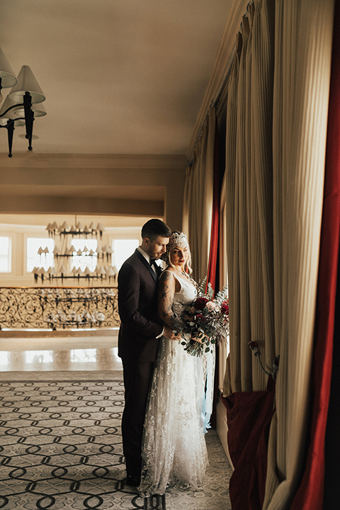 Dana point outdoor wedding shoot at monarch beach resort bride form fitting lace gown with thin straps and plunging neckline with groom burgundy tuxedo with matching vest and black bow tie with floral boutonniere bride holding red and green floral bridal bouquet