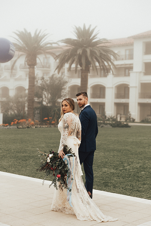 Dana point outdoor wedding shoot at monarch beach resort bride form fitting lace gown with long sleeves and high neckline with groom navy blue suit with matching vest and white dress shirt with long blush pink striped tie and matching pocket square walking and holding hands