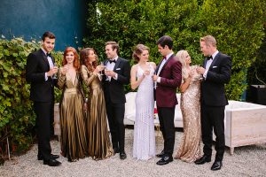 Los angeles glamorous wedding shoot at the fig house bride form fitting light blue beaded gown with thin halter straps and leg slit with groom burgundy tuxedo coat with black tuxedo pants and white dress shirt with burgundy bow tie with bridesmaids long gold dresses and groom black notch lapel tuxedos with black bow ties standing and holding drinks