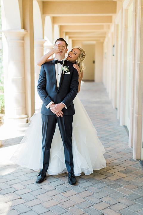 Laguna beach wedding bride ball gown with thin spaghetti straps and a sweetheart neckline with groom navy blue tuxedo with black shawl lapel and white dress shirt with black bow tie and white floral boutonniere bride behind groom covering eyes for first look