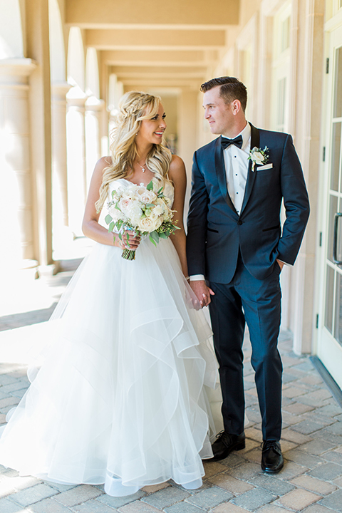Laguna beach wedding bride ball gown with thin spaghetti straps and a sweetheart neckline with groom navy blue tuxedo with black shawl lapel and white dress shirt with black bow tie and white floral boutonniere holding hands and bride holding white and green floral bridal bouquet