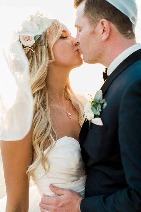 Laguna beach wedding bride ball gown with thin spaghetti straps and a sweetheart neckline with groom navy blue tuxedo with black shawl lapel and white dress shirt with black bow tie and white floral boutonniere kissing