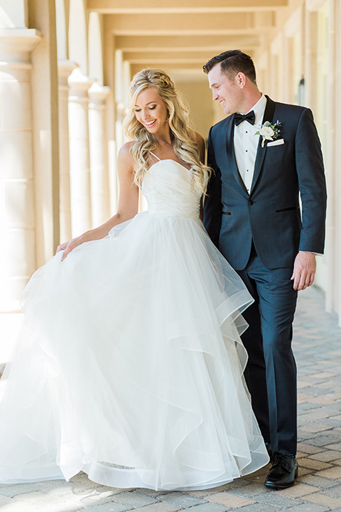 Laguna beach wedding bride ball gown with thin spaghetti straps and a sweetheart neckline with groom navy blue tuxedo with black shawl lapel and white dress shirt with black bow tie and white floral boutonniere hugging and bride holding dress