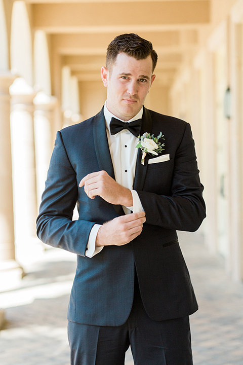 Laguna beach wedding groom navy blue tuxedo with a black shawl lapel and white dress shirt with black bow tie and white pocket square with white floral boutonniere holding jacket sleeve