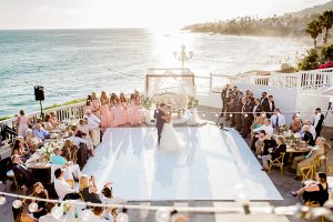 Laguna beach wedding bride ball gown with thin spaghetti straps and a sweetheart neckline with groom navy blue tuxedo with black shawl lapel and white dress shirt with black bow tie and white floral boutonniere first dance with ocean view and hanging lights