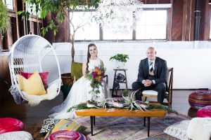 Santa barbara greenery wedding shoot at the narrative loft bride two piece gown with high neckline crop top and tulle skirt with pink and yellow floral bridal bouquet with groom grey suit with matching vest and white dress shirt with long striped tie and floral boutonniere sitting on chairs