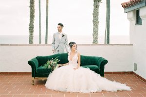 Orange county wedding shoot at ole hanson beach club bride ball gown with lace bodice and straps with plunging neckline halter design with crystal headpiece and groom light grey suit with matching vest and white dress shirt with long ivory tie with white and purple floral boutonniere sitting on couch