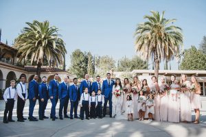 Orange county wedding at the colony house bride long sleeve lace gown with big sleeves and plunging neckline with sheer details with groom cobalt blue suit with matching vest and white dress shirt with long black skinny tie and white floral boutonniere with bridesmaids long blush pink dresses with groomsmen cobalt blue suits with long black skinny ties