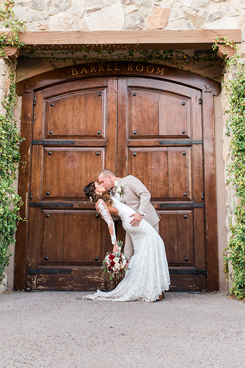 Temecula outdoor wedding at ponte winery bride form fitting lace gown with long sleeves and illusion open back design with sweetheart neckline and groom tan suit with matching vest and white dress shirt with matching tan bow tie and white floral boutonniere kissing bride holding white and red floral bridal bouquet