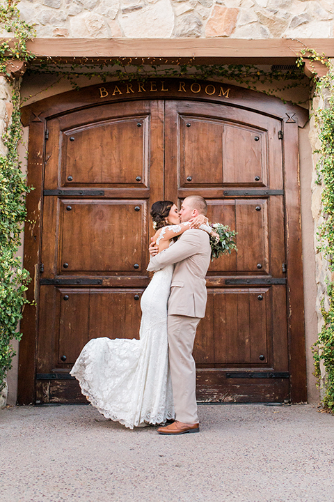 Temecula outdoor wedding at ponte winery bride form fitting lace gown with long sleeves and illusion open back design with sweetheart neckline and groom tan suit with matching vest and white dress shirt with matching tan bow tie and white floral boutonniere kissing bride holding white and red floral bridal bouquet