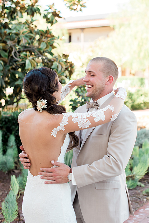 Temecula outdoor wedding at ponte winery bride form fitting lace gown with long sleeves and illusion open back design with sweetheart neckline and groom tan suit with matching vest and white dress shirt with matching tan bow tie and white floral boutonniere hugging first look