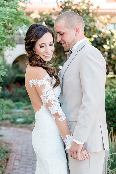 Temecula outdoor wedding at ponte winery bride form fitting lace gown with long sleeves and illusion open back design with sweetheart neckline and groom tan suit with matching vest and white dress shirt with matching tan bow tie and white floral boutonniere holding hands