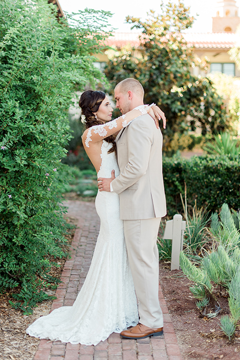 Temecula outdoor wedding at ponte winery bride form fitting lace gown with long sleeves and illusion open back design with sweetheart neckline and groom tan suit with matching vest and white dress shirt with matching tan bow tie and white floral boutonniere hugging