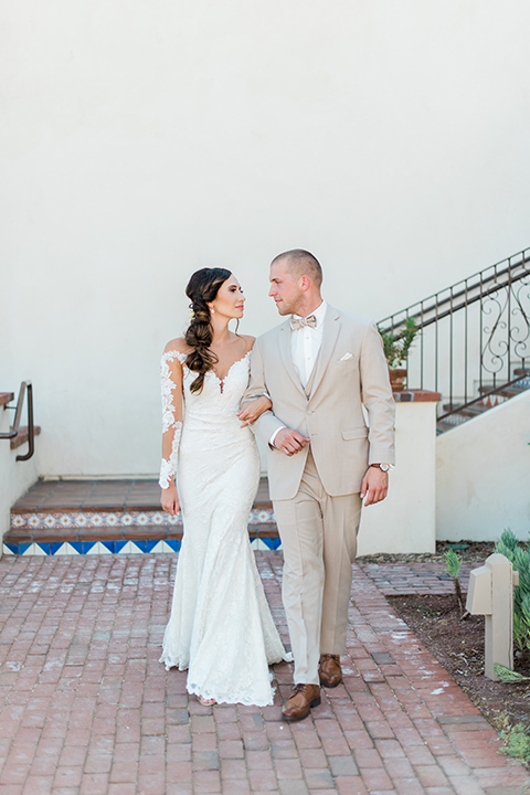 Temecula outdoor wedding at ponte winery bride form fitting lace gown with long sleeves and illusion open back design with sweetheart neckline and groom tan suit with matching vest and white dress shirt with matching tan bow tie and white floral boutonniere bride holding grooms arm