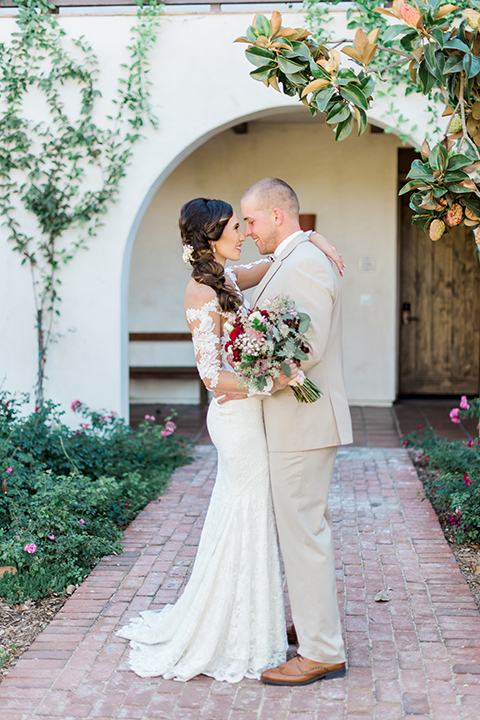 Temecula outdoor wedding at ponte winery bride form fitting lace gown with long sleeves and illusion open back design with sweetheart neckline and groom tan suit with matching vest and white dress shirt with matching tan bow tie and white floral boutonniere hugging bride holding white and red floral bridal bouquet