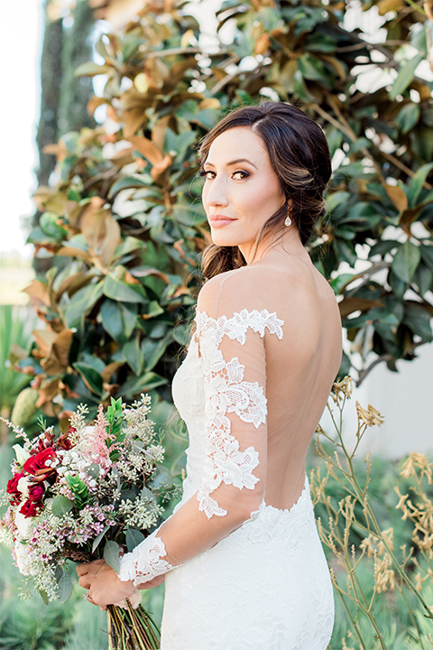Temecula outdoor wedding at ponte winery bride form fitting lace gown with long sleeves and illusion open back design with sweetheart neckline holding white and red floral bridal bouquet