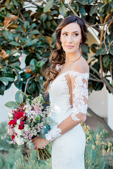 Temecula outdoor wedding at ponte winery bride form fitting lace gown with long sleeves and illusion open back design with sweetheart neckline holding white and red floral bridal bouquet