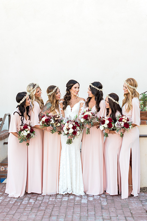 Temecula outdoor wedding at ponte winery bride form fitting lace gown with long sleeves and illusion open back design with sweetheart neckline holding white and red floral bridal bouquet with bridesmaids long blush pink dresses holding white and red floral bridal bouquets with flower crowns