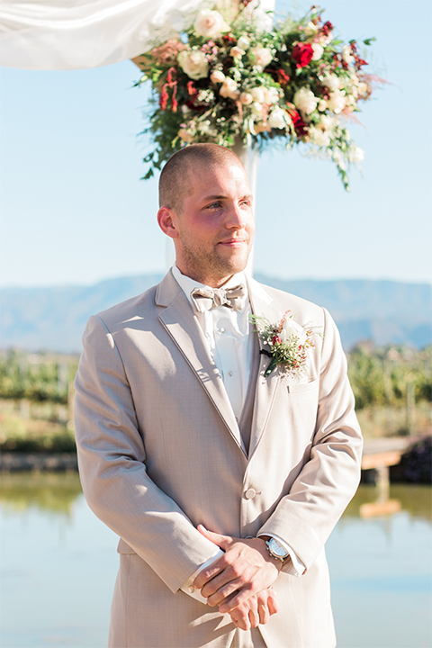 Temecula outdoor wedding at ponte winery groom tan suit with matching vest and white dress shirt with matching tan bow tie with white floral boutonniere watching bride walking down the aisle wedding photo idea for groom