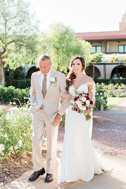 Temecula outdoor wedding at ponte winery bride form fitting lace gown with long sleeves and illusion open back design with sweetheart neckline holding white and red floral bridal bouquet walking down the aisle with dad tan suit with long brown tie