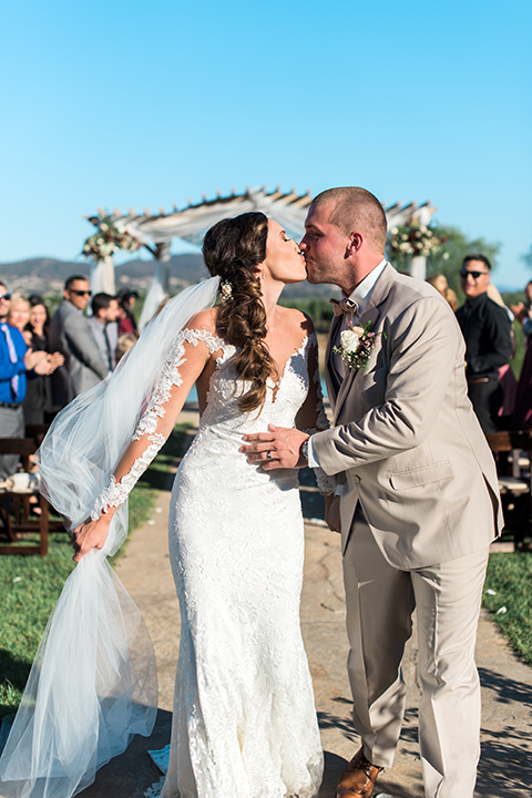 Temecula outdoor wedding at ponte winery bride form fitting lace gown with long sleeves and illusion open back design with sweetheart neckline and groom tan suit with matching vest and white dress shirt with matching tan bow tie and white floral boutonniere kissing in aisle after ceremony