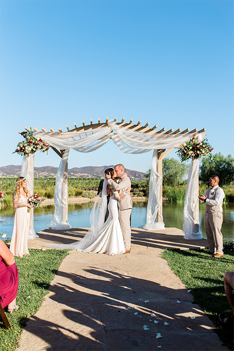 Temecula outdoor wedding at ponte winery bride form fitting lace gown with long sleeves and illusion open back design with sweetheart neckline and groom tan suit with matching vest and white dress shirt with matching tan bow tie and white floral boutonniere kissing during ceremony