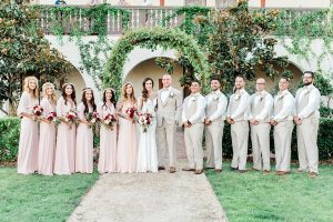 Temecula outdoor wedding at ponte winery bride form fitting lace gown with long sleeves and illusion open back design with sweetheart neckline and groom tan suit with matching vest and white dress shirt with matching tan bow tie and white floral boutonniere with bridesmaids long blush pink dresses with white and red floral bridal bouquets and groomsmen tan suits with matching vests and tan bow ties