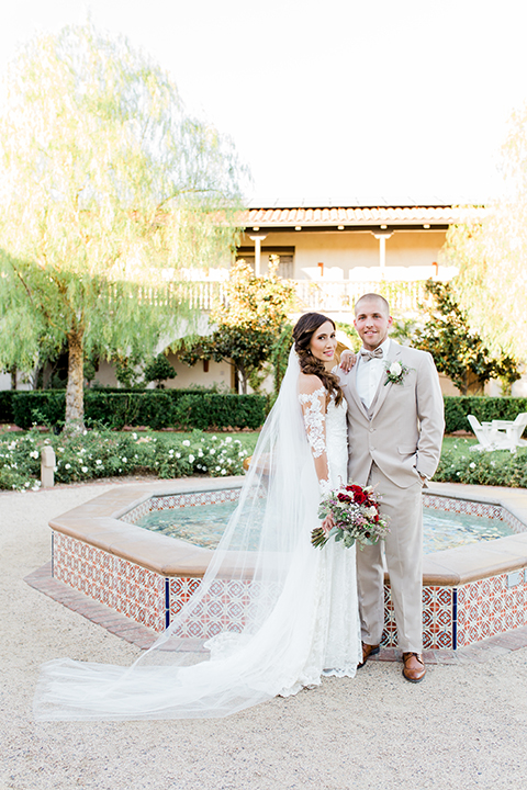 Temecula outdoor wedding at ponte winery bride form fitting lace gown with long sleeves and illusion open back design with sweetheart neckline and groom tan suit with matching vest and white dress shirt with matching tan bow tie and white floral boutonniere standing by fountain bride holding white and red floral bridal bouquet