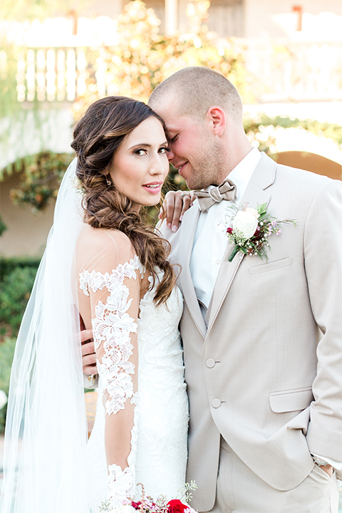 Temecula outdoor wedding at ponte winery bride form fitting lace gown with long sleeves and illusion open back design with sweetheart neckline and groom tan suit with matching vest and white dress shirt with matching tan bow tie and white floral boutonniere hugging bride holding white and red floral bridal bouquet