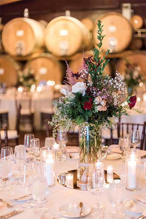 Temecula outdoor wedding at ponte winery reception set up with white table linen and dark chairs with white and green flower centerpiece decor with wine barrels along walls and gold candle decor with wine glasses