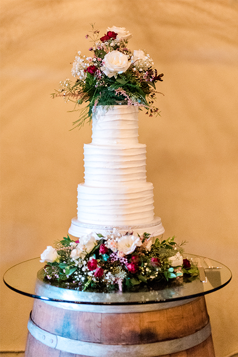 Temecula outdoor wedding at ponte winery reception set up with white table linen and dark chairs with white and green flower centerpiece decor with wine barrels along walls and gold candle decor with wine glasses wine barrel with clear glass top and four tier white wedding cake with white and red and green floral decor