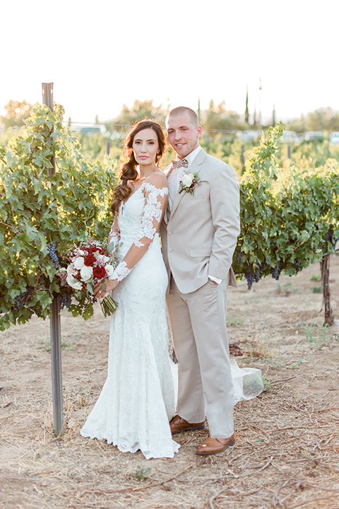Temecula outdoor wedding at ponte winery bride form fitting lace gown with long sleeves and illusion open back design with sweetheart neckline and groom tan suit with matching vest and white dress shirt with matching tan bow tie and white floral boutonniere bride holding white and red floral bridal bouquet