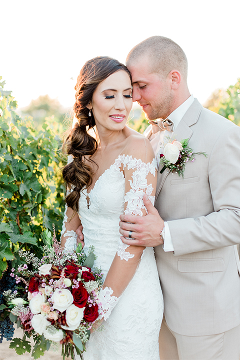 Temecula outdoor wedding at ponte winery bride form fitting lace gown with long sleeves and illusion open back design with sweetheart neckline and groom tan suit with matching vest and white dress shirt with matching tan bow tie and white floral boutonniere hugging bride holding white and red floral bridal bouquet