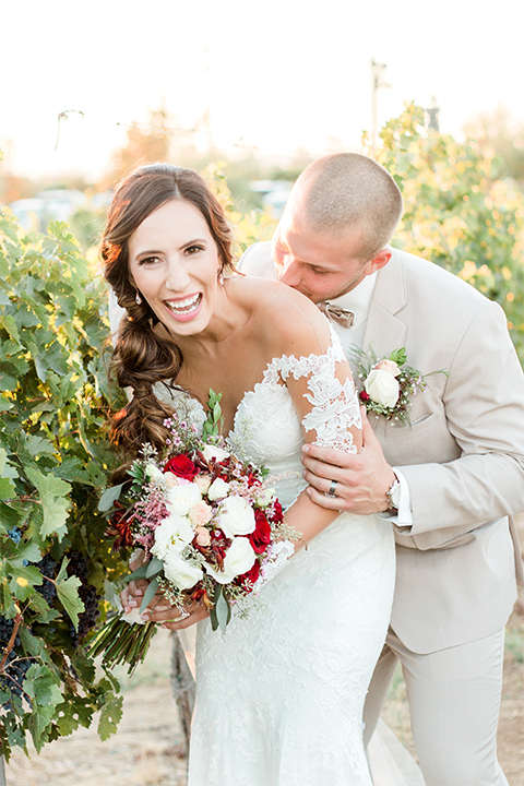 Temecula outdoor wedding at ponte winery bride form fitting lace gown with long sleeves and illusion open back design with sweetheart neckline and groom tan suit with matching vest and white dress shirt with matching tan bow tie and white floral boutonniere laughing bride holding white and red floral bridal bouquet