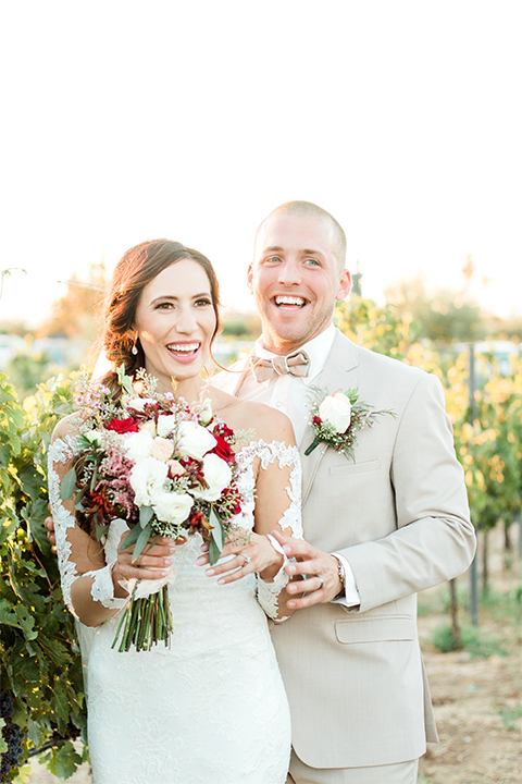 Temecula outdoor wedding at ponte winery bride form fitting lace gown with long sleeves and illusion open back design with sweetheart neckline and groom tan suit with matching vest and white dress shirt with matching tan bow tie and white floral boutonniere bride holding white and red floral bridal bouquet