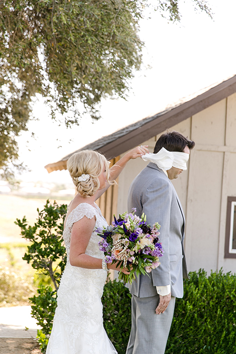 Temecula outdoor wedding at callaway winery bride form fitting lace gown with thick straps and open back design with plunging neckline with groom heather grey suit with matching vest and white dress shirt with long ivory tie and purple floral boutonniere first look unwrapping blindfold