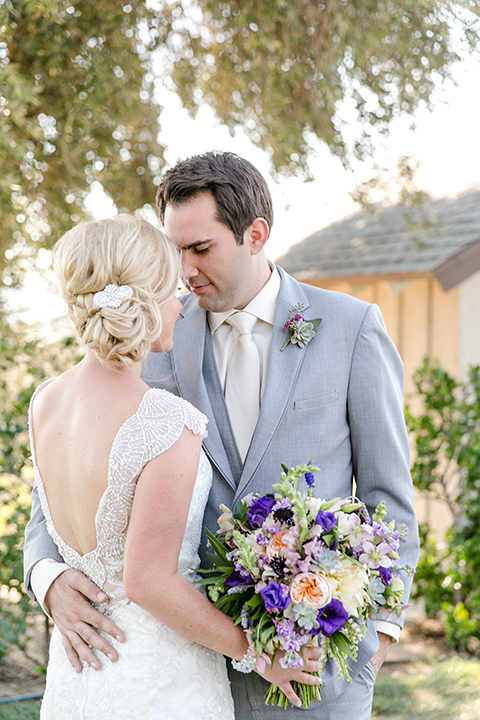 Temecula outdoor wedding at callaway winery bride form fitting lace gown with thick straps and open back design with plunging neckline with groom heather grey suit with matching vest and white dress shirt with long ivory tie and purple floral boutonniere hugging bride holding white and purple floral bridal bouquet