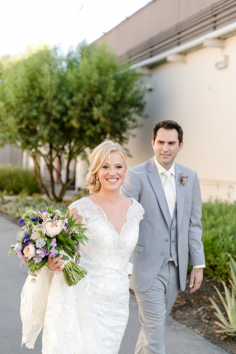 Temecula outdoor wedding at callaway winery bride form fitting lace gown with thick straps and open back design with plunging neckline with groom heather grey suit with matching vest and white dress shirt with long ivory tie and purple floral boutonniere holding hands bride holding white and purple floral bridal bouquet