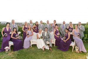 Temecula outdoor wedding at callaway winery bride form fitting lace gown with thick straps and open back design with plunging neckline with groom heather grey suit with matching vest and white dress shirt with long ivory tie and purple floral boutonniere sitting down with wedding party bridesmaids long purple dresses and groomsmen heather grey suits with long purple ties