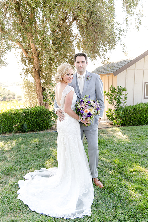 Temecula outdoor wedding at callaway winery bride form fitting lace gown with thick straps and open back design with plunging neckline with groom heather grey suit with matching vest and white dress shirt with long ivory tie and purple floral boutonniere hugging bride holding white and purple floral bridal bouquet