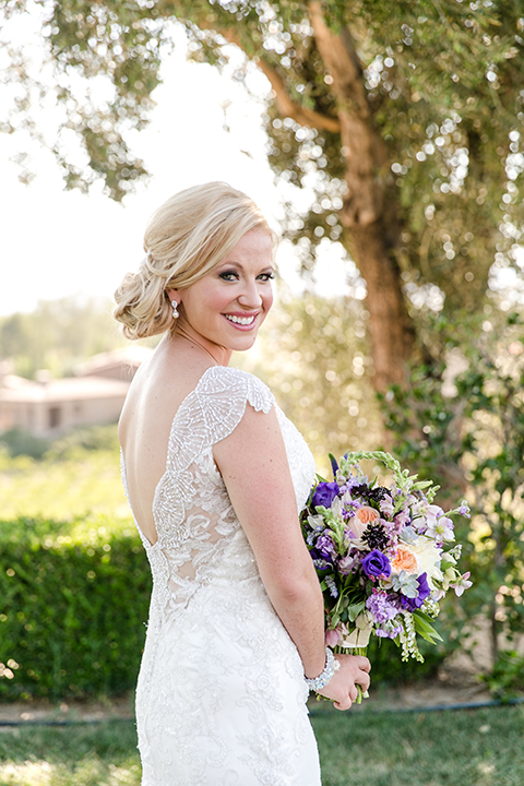 Temecula outdoor wedding at callaway winery bride form fitting lace gown with thick straps and open back design with plunging neckline with white and purple floral bridal bouquet