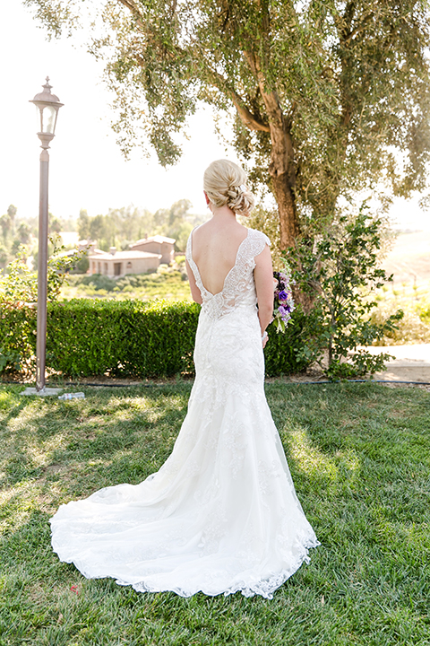 Temecula outdoor wedding at callaway winery bride form fitting lace gown with thick straps and open back design with plunging neckline with white and purple floral bridal bouquet