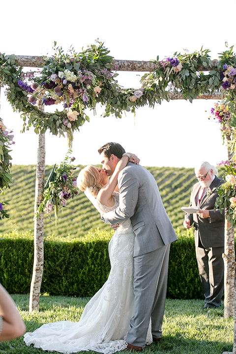 Temecula outdoor wedding at callaway winery bride form fitting lace gown with thick straps and open back design with plunging neckline with groom heather grey suit with matching vest and white dress shirt with long ivory tie and purple floral boutonniere kissing during ceremony