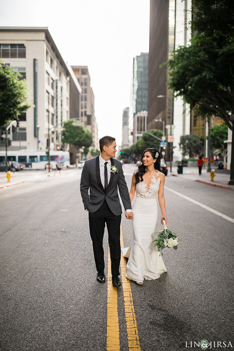 Downtown los angeles wedding bride form fitting lace gown with illusion back and high neckline with crystal hair piece with groom charcoal grey tuxedo with black shawl lapel and white dress shirt with long black skinny tie and white floral boutonniere walking in the street holding hands